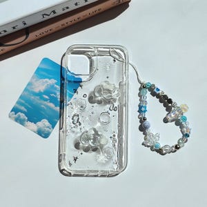 May include: Clear phone case with cloud and star embellishments. The case has a beaded wrist strap with blue, white, and iridescent beads. A card with a sky and cloud image is next to the case.