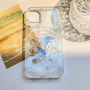 Può includere: Custodia trasparente per telefono con design liquido e glitter. La custodia presenta accenti blu e oro, con piccoli elementi decorativi. La custodia è progettata per proteggere il telefono aggiungendo un tocco decorativo.