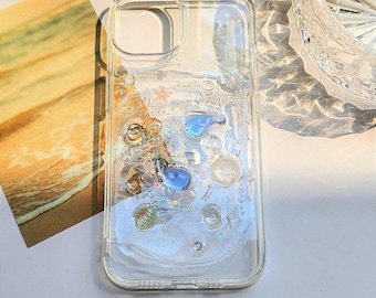 May include: A clear, transparent phone case featuring a liquid-filled back panel containing floating blue whale-shaped charms, small iridescent glitter particles, and various clear, spherical beads that move within the sealed compartment.