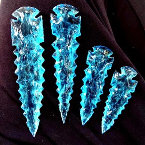 May include: Four blue glass arrowheads of different sizes. Each arrowhead has a jagged edge and a notched top. The glass is translucent, with white and blue swirls. The arrowheads are displayed against a dark background.