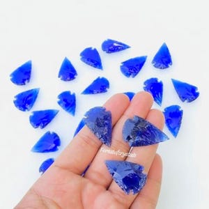 Può includere: Una collezione di pezzi di vetro blu brillante a forma di punta di freccia. Alcuni sono tenuti in una mano, mentre altri sono sparsi su una superficie bianca. Il testo "Forestofcrystals" è visibile sulla mano.