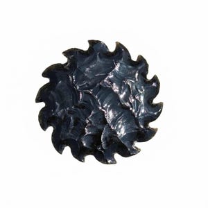 Puede incluir: Una piedra de obsidiana circular con una superficie negra brillante y un borde dentado en forma de engranaje. La piedra presenta una serie de patrones de escamas afiladas y superpuestas que crean una apariencia texturizada y reflectante.