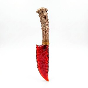 May include: A decorative knife featuring a translucent, vibrant red blade with a jagged, textured edge. The handle is sculpted to resemble a piece of weathered, light brown wood or bone, featuring intricate organic ridges and a wrapped band.
