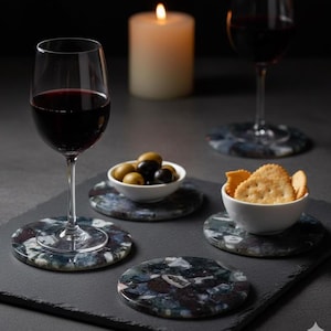 May include: A sophisticated table arrangement with glasses of red wine, bowls of olives and crackers, and patterned round coasters. A lit candle adds a warm glow. The coasters have a dark, mottled design, adding to the elegant setting.
