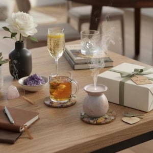 May include: A still life arrangement on a wooden table features a white peony in a black vase, a glass of champagne, and a cup of tea. A small pink essential oil diffuser emits vapor. Other items include a gift box and a notebook.