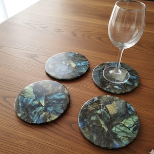 May include: Four circular coasters with a distinctive, shimmering pattern of blues, greens, and yellows, reminiscent of natural stone. A wine glass sits on one coaster. The coasters are on a wooden surface.