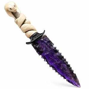 Puede incluir: Una daga decorativa con una hoja de vidrio púrpura translúcido de borde dentado y tallado. El mango está hecho de un material blanco hueso esculpido con la forma de una cobra enroscada, con una guarda de metal negro que separa la hoja del mango.