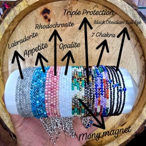Op de afbeelding: Een verzameling kralenarmbanden in verschillende kleuren, waaronder grijs, blauw, roze en zwart, gepresenteerd op een witte cilindrische standaard. De armbanden zijn gelabeld met teksten als "Labradorite", "Rhodochrosite" en "Black Obsidian Evil Eye."
