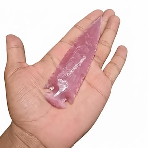 May include: A pink arrowhead made of stone, held in a hand. The arrowhead has a pointed tip and a notched base. The text "Forestofcrystals" is printed on the arrowhead.