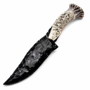 May include: A fixed-blade knife featuring a sharp, glossy black obsidian blade with a chipped, textured edge. The handle is crafted from a light-colored, textured antler material, intricately carved with detailed scrollwork patterns throughout.