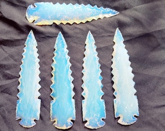 May include: Four opalescent glass arrowheads with a blue and white color scheme are displayed on a dark gray surface. One arrowhead is positioned above the others. The arrowheads have a jagged edge design.