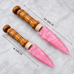 Puede incluir: Dos dagas decorativas con hojas de color rosa translúcido y mangos de madera marrón pulida con ranuras horizontales. La daga más grande mide 25,4 cm de largo y la más pequeña mide 20,32 cm de largo.