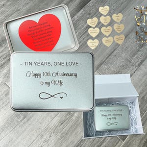 May include: A silver metal tin box with the text "TIN YEARS, ONE LOVE ~ Happy 10th Anniversary to my Wife" and an infinity symbol. Inside is a red heart-shaped card with printed text and ten small wooden heart tokens with engraved phrases.