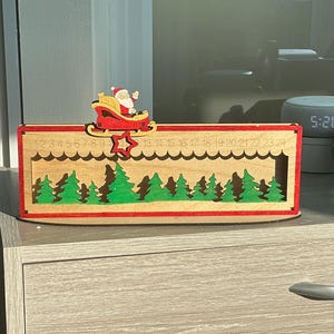 May include: A rectangular wooden Christmas advent calendar featuring a red frame and a natural wood interior. It displays a green forest silhouette, a red star, and a Santa Claus in a sleigh. The numbers 1 through 24 are engraved along the top edge.