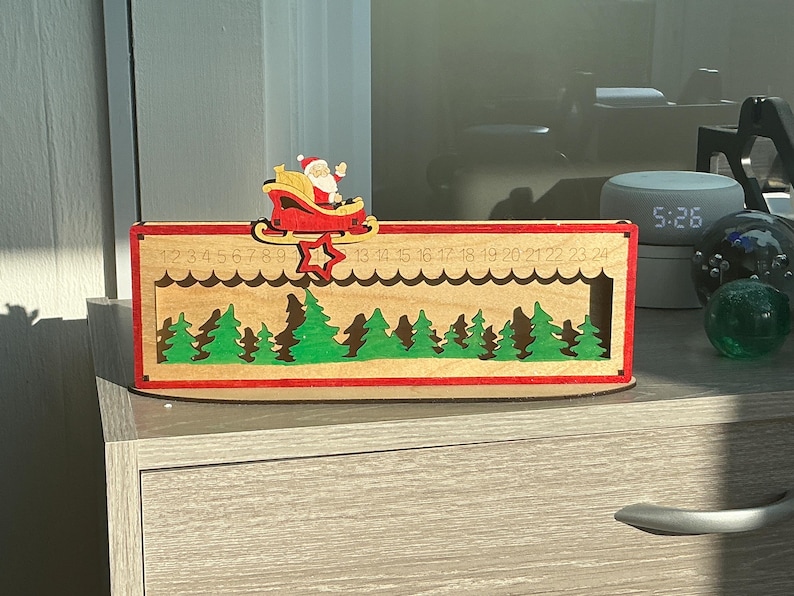 May include: A rectangular wooden Christmas advent calendar featuring a red frame and a natural wood interior. It displays a green forest silhouette, a red star, and a Santa Claus in a sleigh. The numbers 1 through 24 are engraved along the top edge.