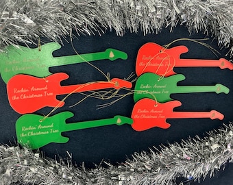 May include: Red and green guitar-shaped Christmas tree ornaments. Each ornament has the text "Rockin' Around the Christmas Tree" printed on it. The ornaments are strung with gold string and surrounded by silver tinsel.