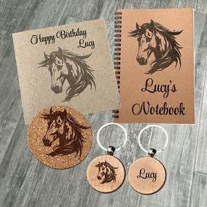 May include: A set of horse-themed gifts, including a notebook, coaster, and keyrings. The notebook and coaster are brown with a horse head design and the text "Lucy's Notebook" and "Happy Birthday Lucy". The keyrings are wooden with a horse head design and the name "Lucy".