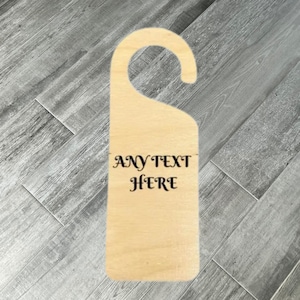 May include: A light-coloured wooden door hanger with a curved top and rectangular body. The text "ANY TEXT HERE" is printed in black, stylized font. The hanger is designed to be hung on a door knob.