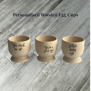 May include: Three personalised wooden egg cups. Each cup is light-coloured with a rounded bowl shape and a pedestal base. The cups are engraved with the phrases "You crack me up!", "You're a good egg!", and "Billy's dippy egg!"