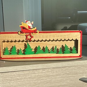 May include: A rectangular wooden Christmas advent calendar featuring a laser-cut forest of green trees. A red sleigh with Santa Claus sits on a sliding track at the top, marked with numbers 1 through 24, and includes a red star pointer.