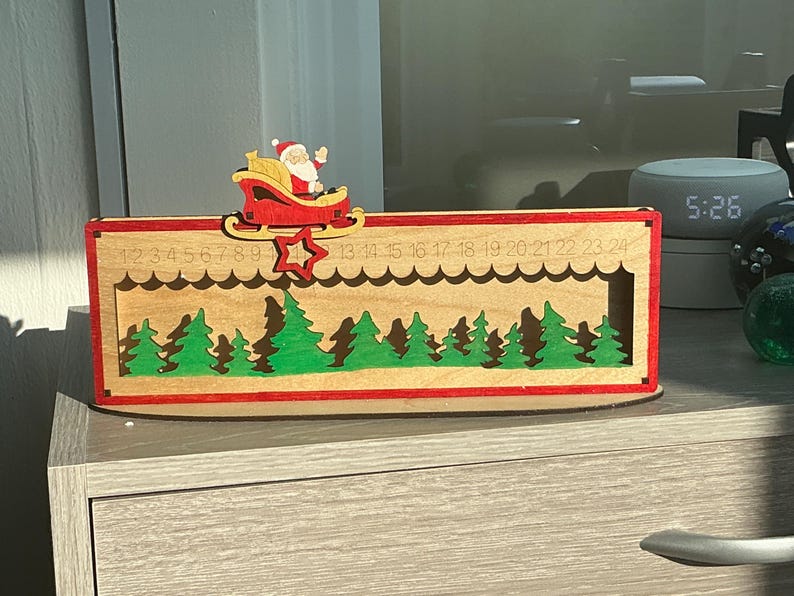 May include: A rectangular wooden Christmas advent calendar featuring a laser-cut forest of green trees. A red sleigh with Santa Claus sits on a sliding track at the top, marked with numbers 1 through 24, and includes a red star pointer.