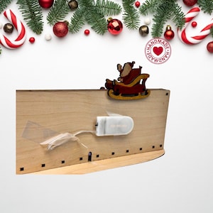 May include: A light-colored wooden decorative piece featuring a cutout of Santa Claus in a red sleigh attached to the top edge. A white plastic battery pack is secured to the back with clear tape, connected by a thin wire to the wooden structure.