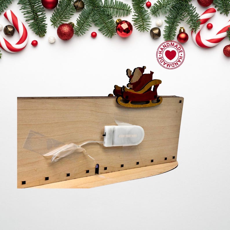 May include: A light-colored wooden decorative piece featuring a cutout of Santa Claus in a red sleigh attached to the top edge. A white plastic battery pack is secured to the back with clear tape, connected by a thin wire to the wooden structure.