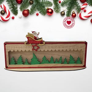 May include: A rectangular wooden Christmas advent calendar featuring a laser-cut Santa in a sleigh, a red star marker, and numbers 1 through 24 engraved along the top. The base includes a green forest silhouette against a light wood background.