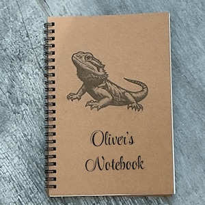 May include: Spiral-bound notebook with a brown cover featuring a detailed illustration of a lizard. The text "Oliver's Notebook" is printed in black script below the lizard image. The notebook has a black spiral binding.