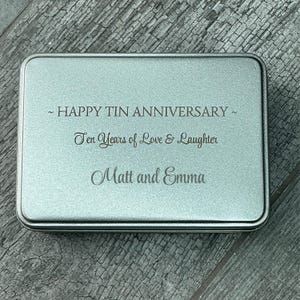 May include: A rectangular, silver-coloured tin with rounded corners. The text "~ HAPPY TIN ANNIVERSARY ~ Ten Years of Love & Laughter Matt and Emma" is engraved on the lid. A gift for a ten-year anniversary.