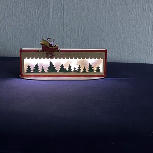 May include: A rectangular wooden light box featuring a cutout silhouette of a forest with green trees and a white scalloped border. A small wooden figure of Santa Claus in a red sleigh is attached to the top edge. The interior emits a warm white glow.