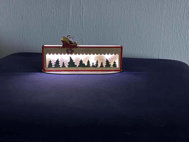 May include: A rectangular wooden light box featuring a cutout silhouette of a forest with green trees and a white scalloped border. A small wooden figure of Santa Claus in a red sleigh is attached to the top edge. The interior emits a warm white glow.