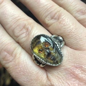 May include: A silver ring with a skull-shaped amber stone set in the center. The stone has two orange gemstones for eyes.