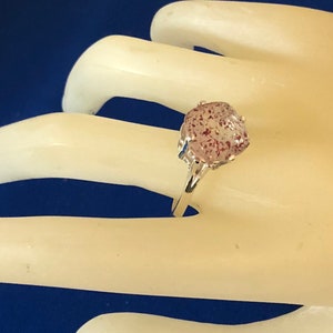 May include: A silver ring featuring a large, oval-shaped gemstone with a speckled, pinkish-red appearance. The ring's band is thin and polished, and the gemstone is held in place by four prongs. The ring is displayed against a white background.