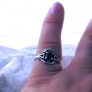 May include: A silver ring with an oval, dark gemstone. The ring has an ornate, filigree design around the stone and on the band. The ring is shown on a finger, with a blurred background.
