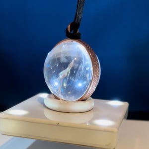 May include: A clear crystal ball pendant with a copper-toned metal frame and a black cord. The pendant is sitting on a white platform with small lights.