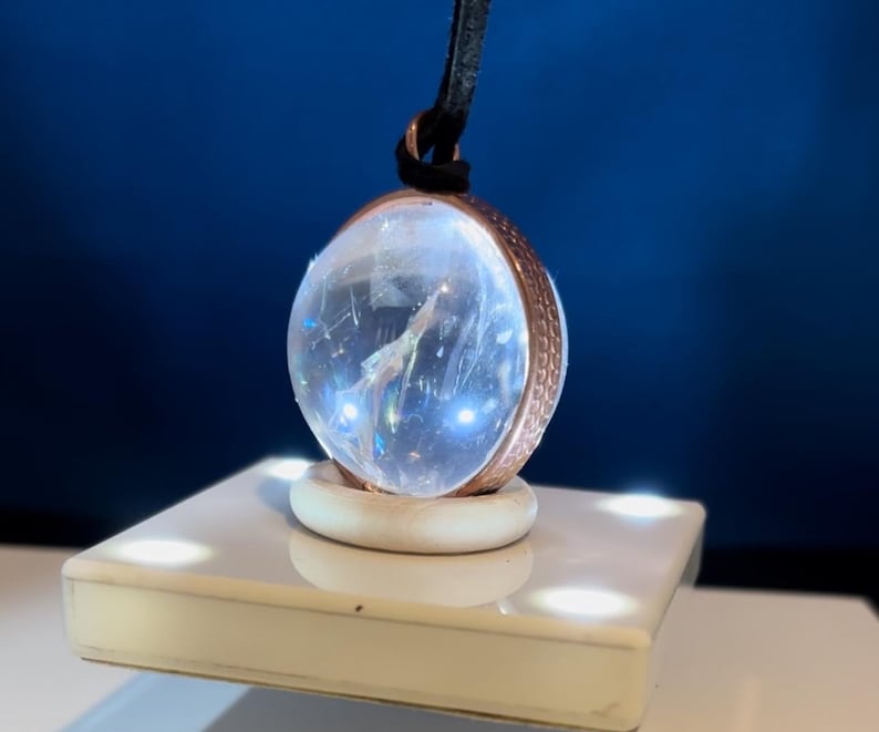 May include: A clear crystal ball pendant with a copper-toned metal frame and a black cord. The pendant is sitting on a white platform with small lights.
