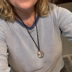 May include: A woman wearing a blue and white striped long-sleeved shirt with a black cord necklace and a round silver pendant.