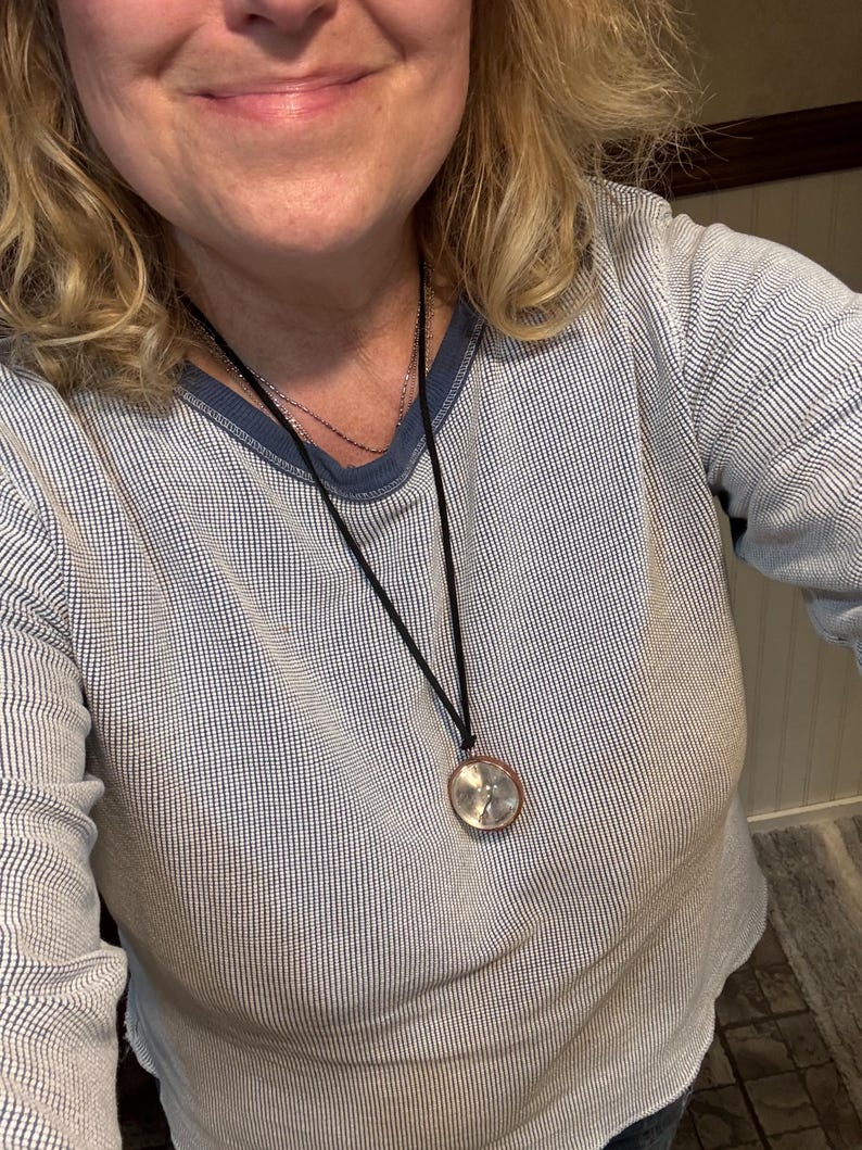 May include: A woman wearing a blue and white striped long-sleeved shirt with a black cord necklace and a round silver pendant.