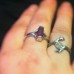 May include: A silver ring with a pink gemstone set in a unique, open design. The ring is worn on a person's finger.