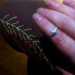 May include: A silver ring with a single round diamond set in a rose-shaped design. The ring is on a finger wearing a brown fabric with white and pink embroidery.