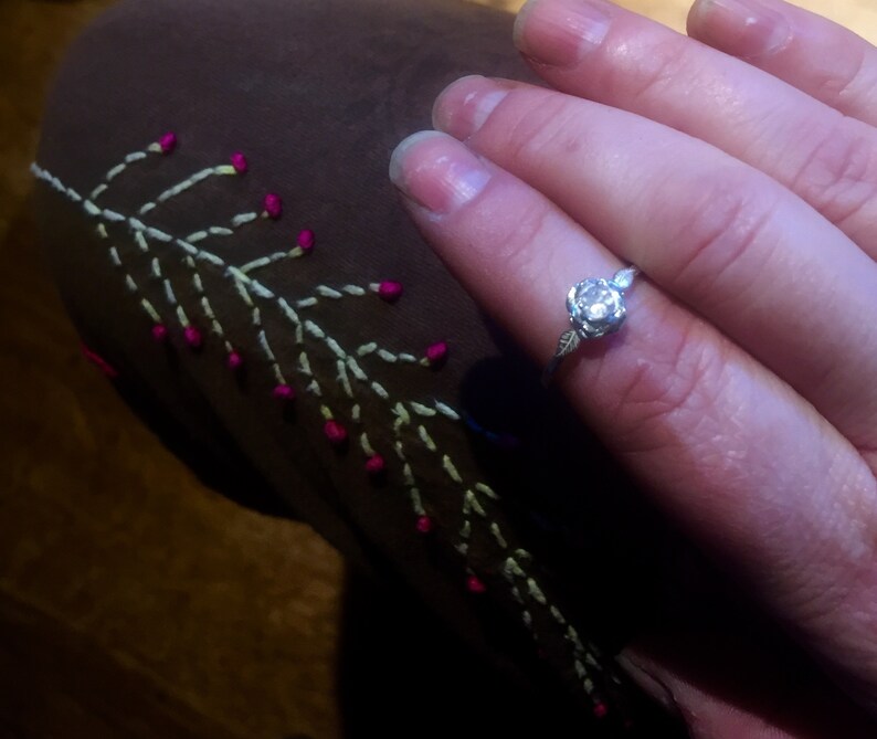 May include: A silver ring with a single round diamond set in a rose-shaped design. The ring is on a finger wearing a brown fabric with white and pink embroidery.