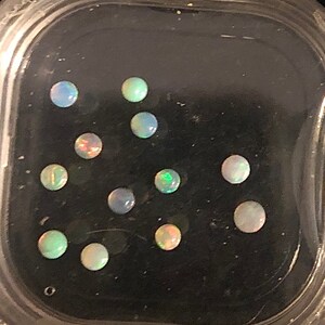 May include: A close-up of a clear plastic container with 11 small, round, iridescent opal gemstones. The gemstones are arranged in a random pattern on a black background.