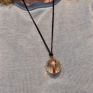 May include: A clear crystal ball pendant with a copper band around the middle, hanging from a black cord necklace.