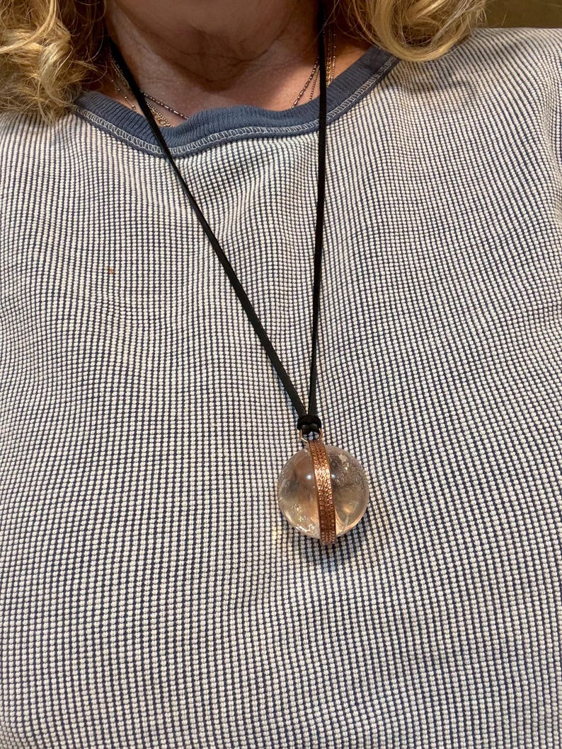 May include: A clear crystal ball pendant with a copper band around the middle, hanging from a black cord necklace.
