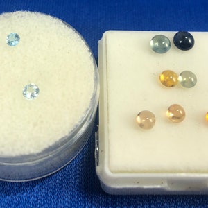 May include: A collection of small, round gemstones in various colors, including blue, green, yellow, and orange. The gemstones are displayed in two small containers.