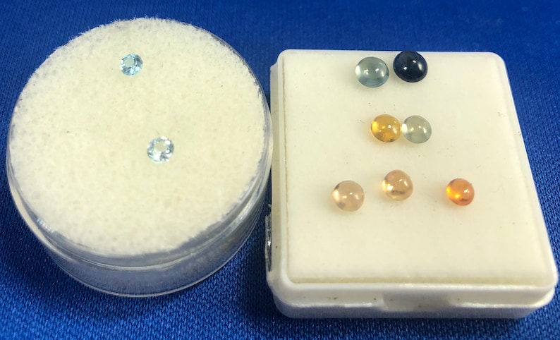 May include: A collection of small, round gemstones in various colors, including blue, green, yellow, and orange. The gemstones are displayed in two small containers.
