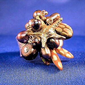 May include: A gold ring with a cluster of dark brown pearls and a gold starfish charm. The ring is set on a blue background.