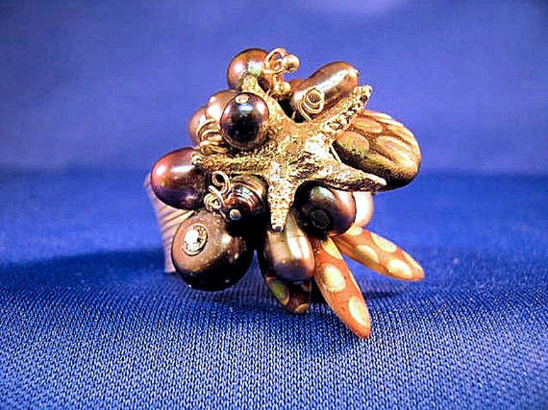 May include: A gold ring with a cluster of dark brown pearls and a gold starfish charm. The ring is set on a blue background.