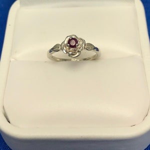 May include: A silver ring with a red gemstone set in a rose design. The ring is in a white ring box.